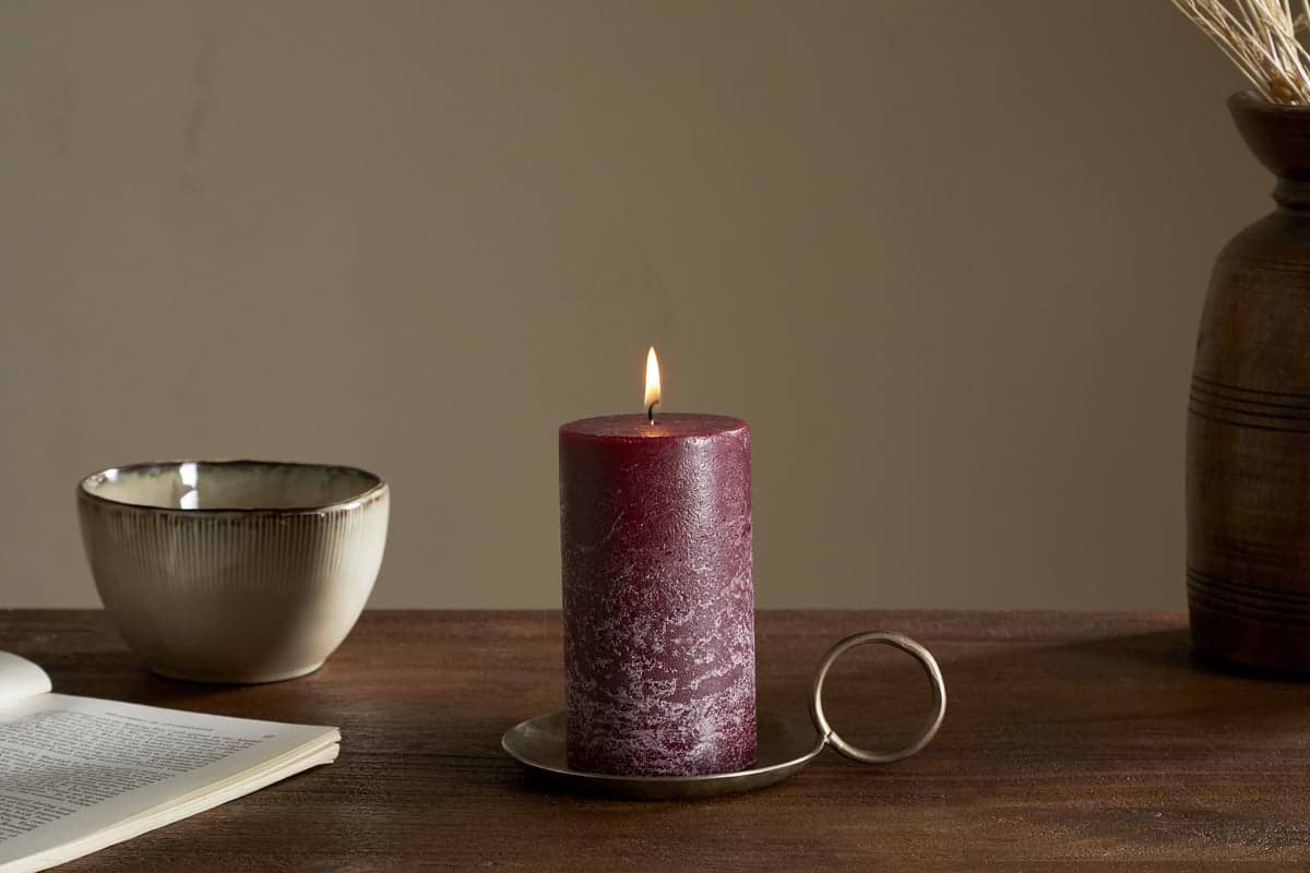 The nkuku Rustic Soy Blend Pillar Candle in Dark Cherry (Small) with a textured finish sits on a metal holder on a wooden table beside an open book, ceramic bowl, and brown vase with dried flowers in soft light.
