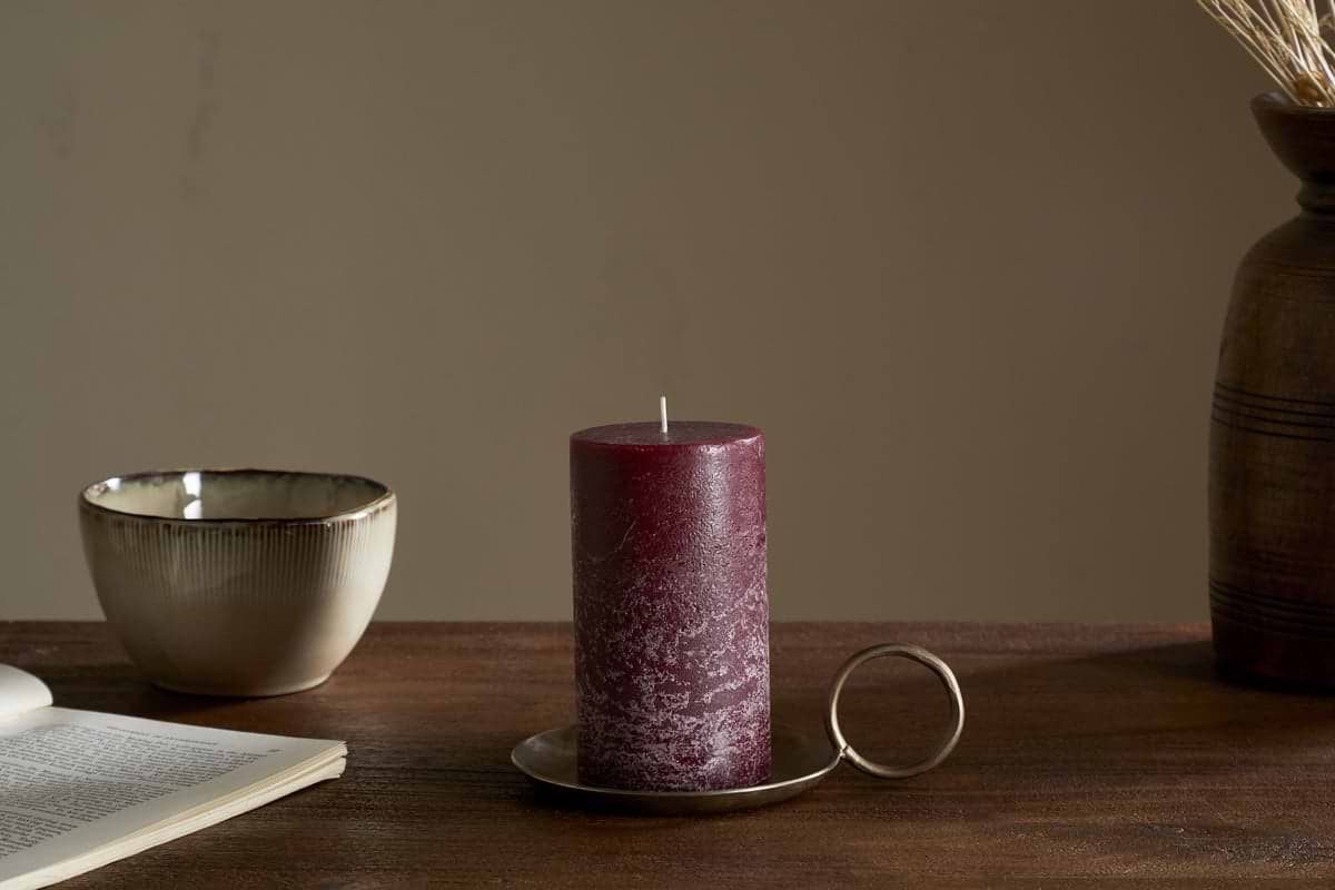 The nkuku Rustic Soy Blend Pillar Candle in Dark Cherry sits on a metal tray with a round handle on a wooden table alongside an open book, ceramic bowl, and brown vase with dried grasses.