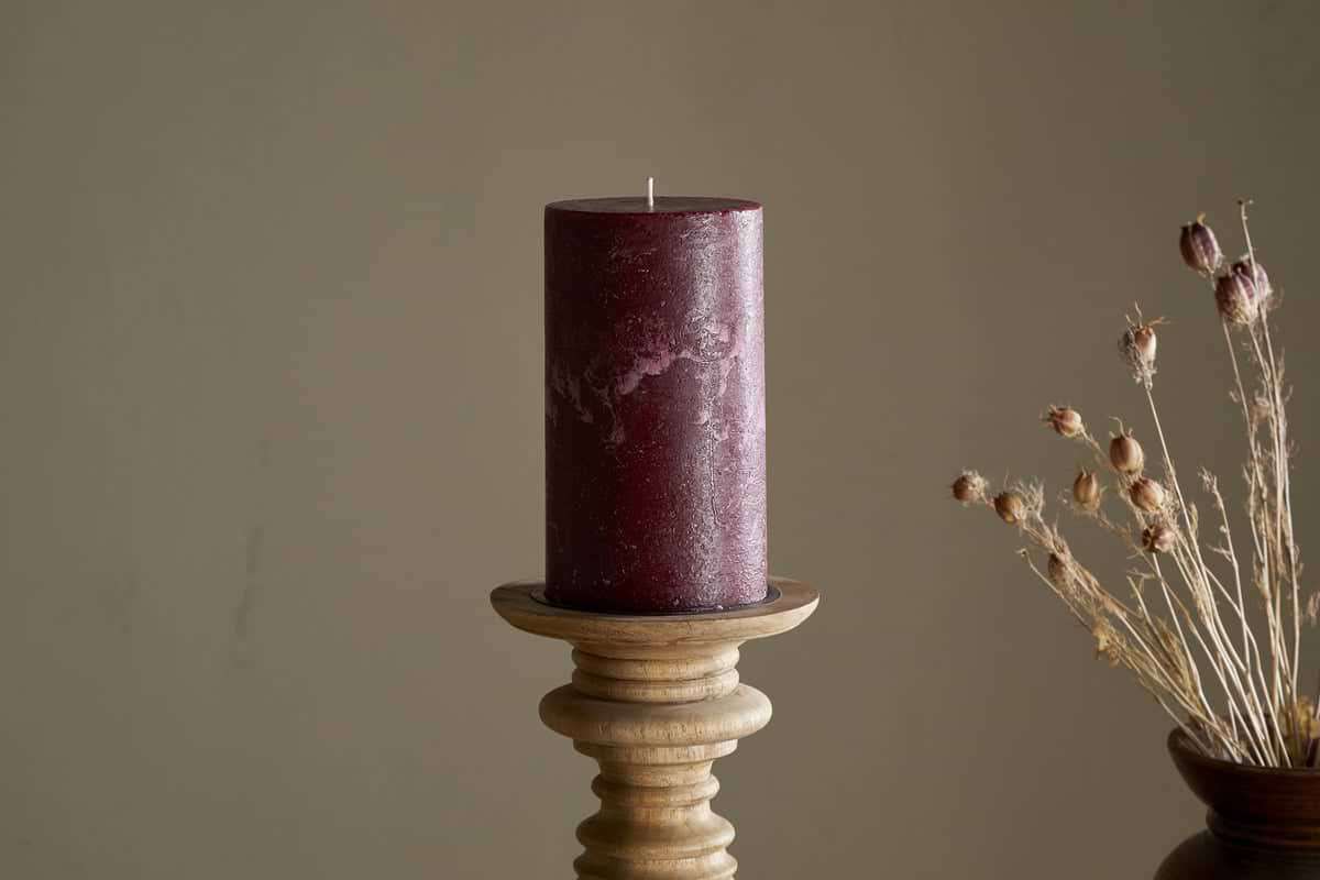 A nkuku Rustic Soy Blend Pillar Candle in Dark Cherry—large, with a textured finish—rests on a wooden holder. To the right, dried flowers in a brown vase contrast against a plain beige wall.