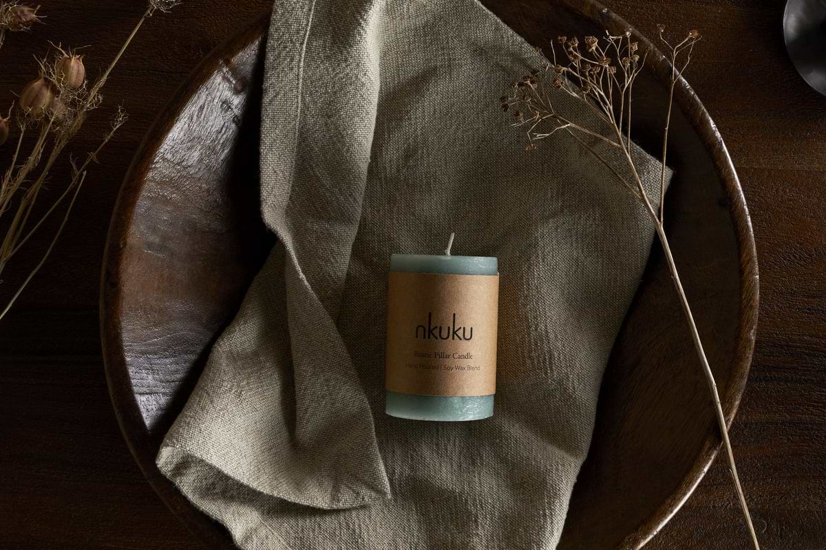 A Rustic Soy Blend Mini Pillar Candle in Sage Green by nkuku, featuring a textured finish, rests on a beige cloth in a wooden bowl with dried plants, atop a dark wooden surface.