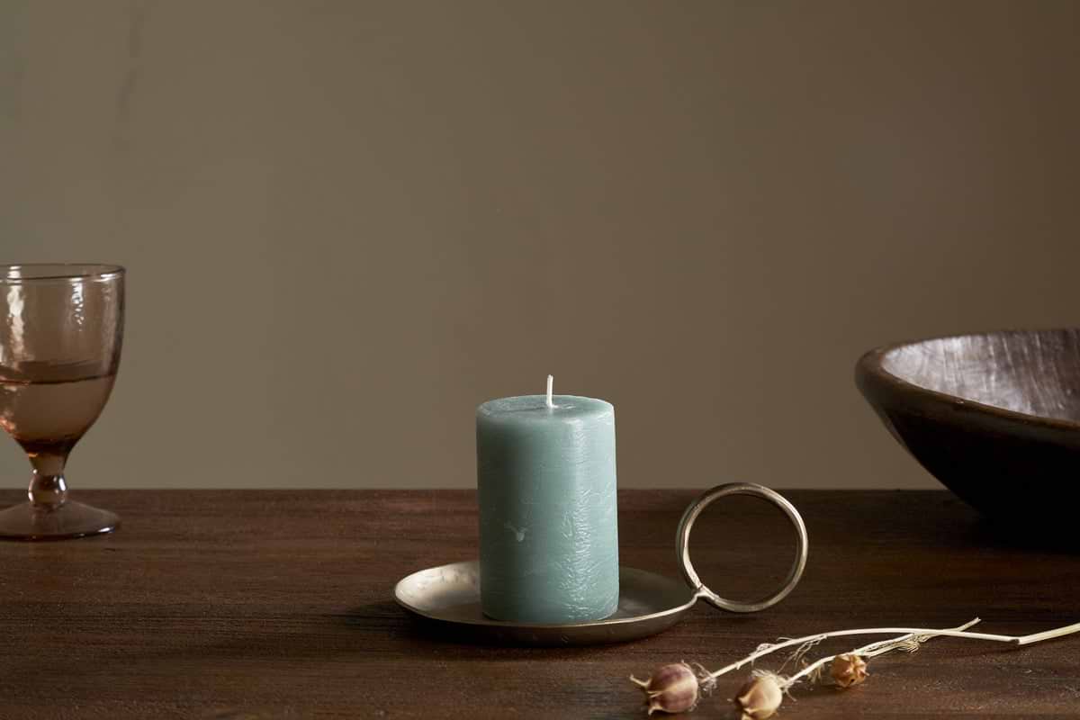 The nkuku Rustic Soy Blend Mini Pillar Candle in Sage Green rests on a brass holder on a wooden table, beside a brown bowl, dried plant, and translucent glass goblet against a neutral backdrop.