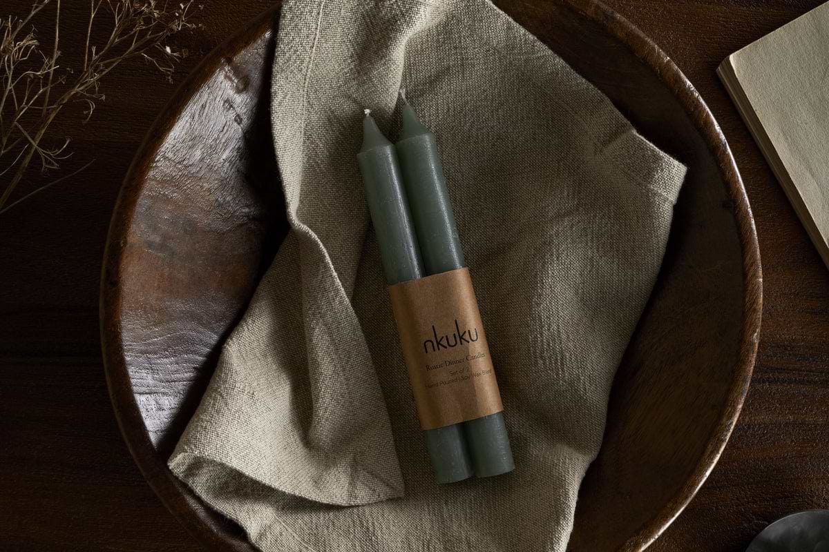 A set of two nkuku Rustic Soy Blend Dinner Candles in sage green, wrapped in a branded label, rest on beige cloth in a wooden bowl, with a dried plant and part of an open book on a dark wooden surface.