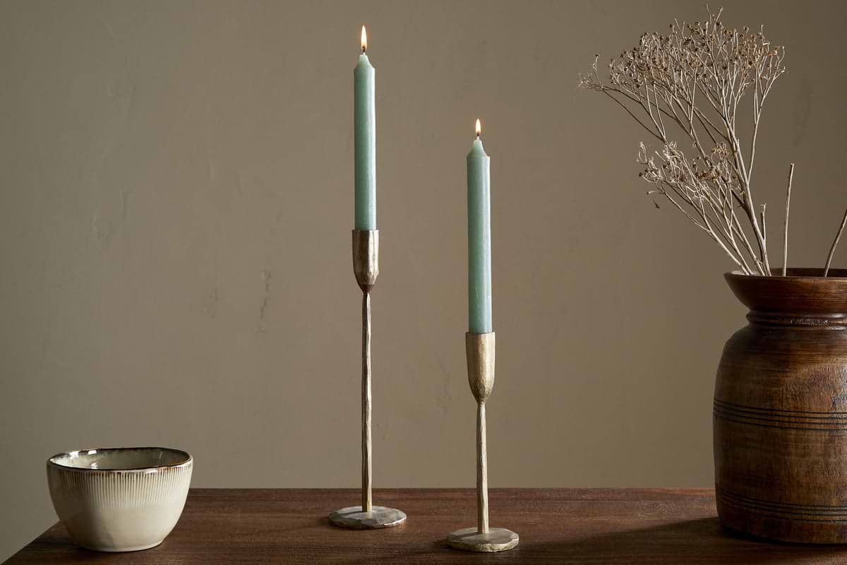 A wooden table features nkukus Rustic Soy Blend Dinner Candle - Sage Green (Set of 2) in brass holders, a ceramic bowl, and a brown vase with dried branches, all set against a beige wall.