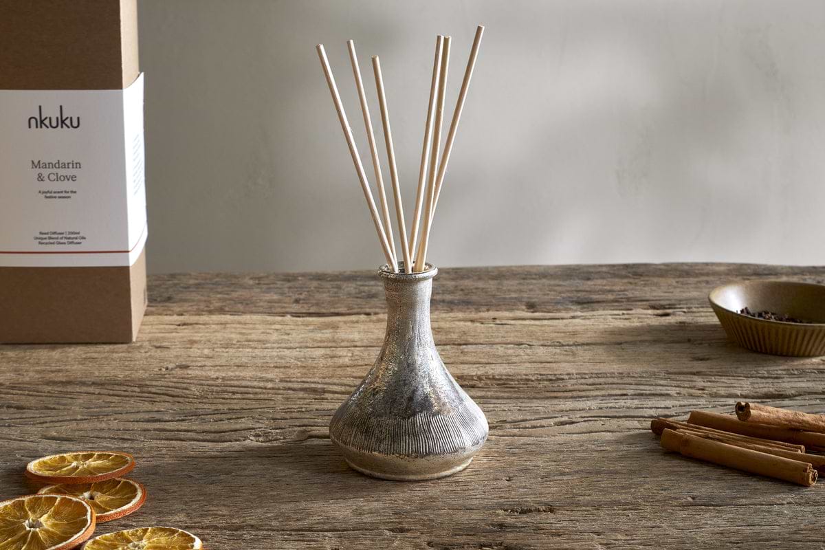 The nkuku Mandarin & Clove Reed Diffuser sits on a wooden table, filling the air with spiced mandarin aroma. Dried orange slices, cinnamon sticks, and cloves surround it, alongside its festive box labeled Mandarin & Clove.