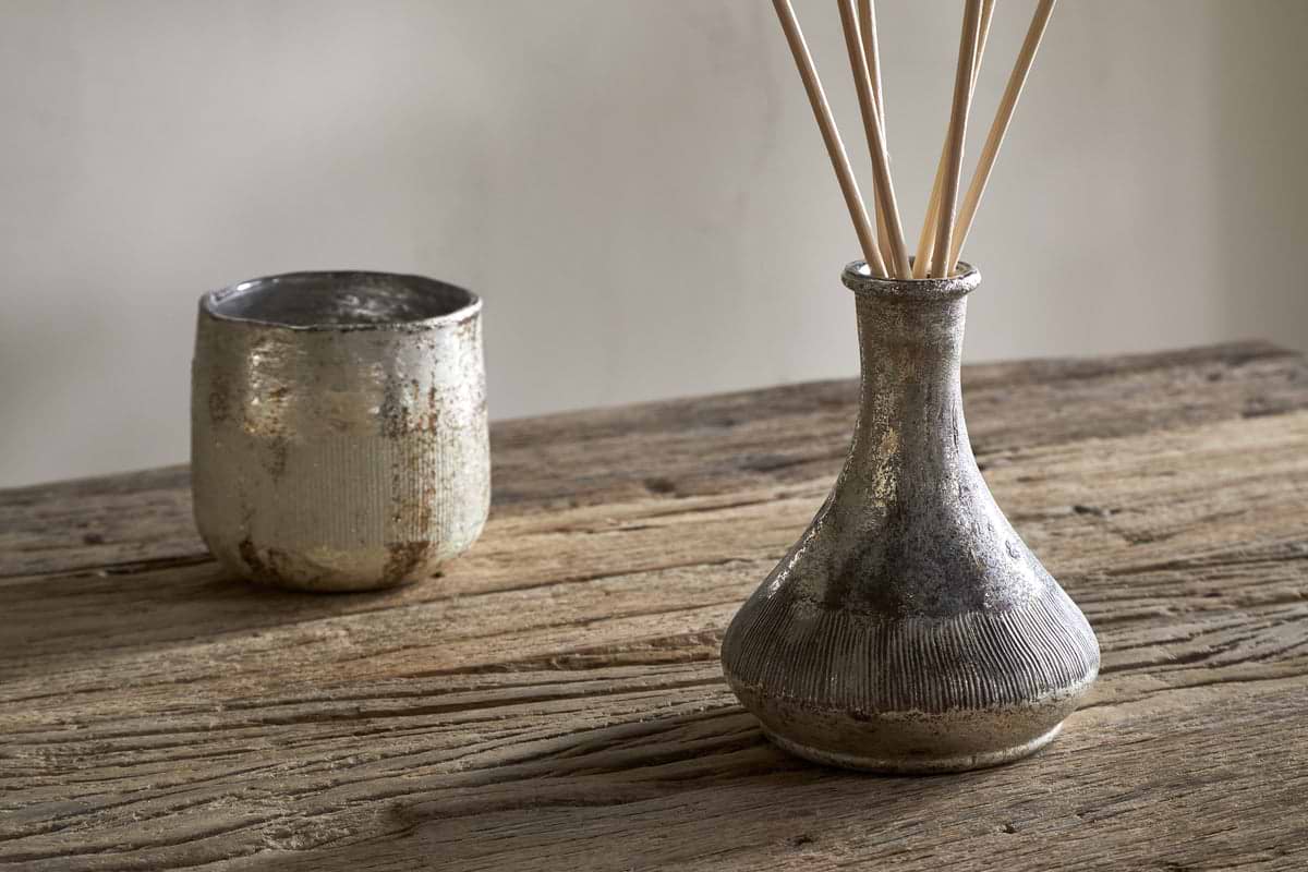 A rustic wooden table displays nkuku’s Mandarin & Clove Reed Diffuser in a textured silver vase, gently filling the air with festive spiced mandarin scent, beside a matching silver cup against a neutral background.