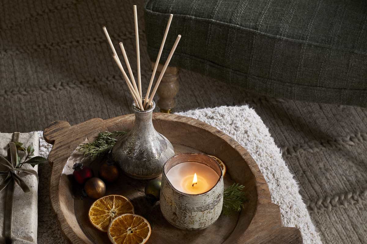 A wooden tray holds a lit candle, dried orange slices, festive ornaments, and the nkuku Mandarin & Clove Reed Diffuser in a vase for a festive scent. The tray rests on a textured white surface beside a green upholstered chair.