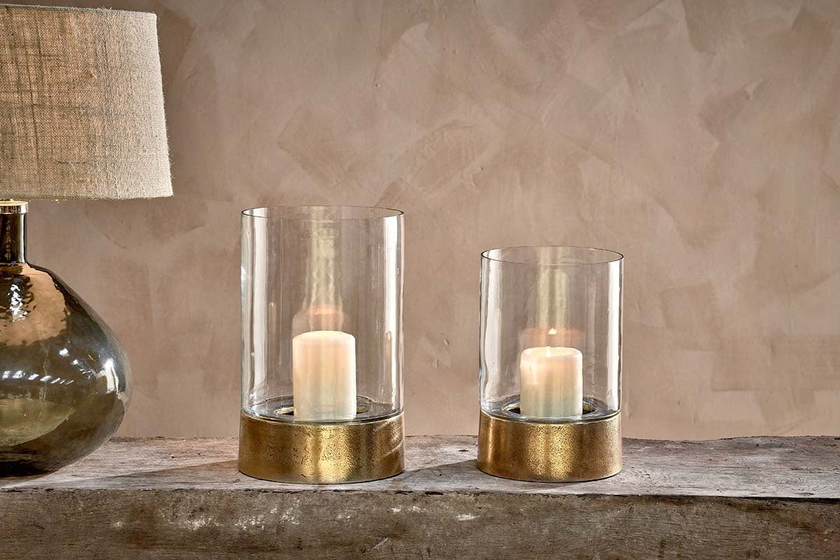 Two Rajura Hurricane Lanterns by nkuku with gold bases rest on a rustic wooden surface beside a glass-based lamp with a textured beige shade, all set against a textured beige wall, creating warm and cozy ambient lighting.