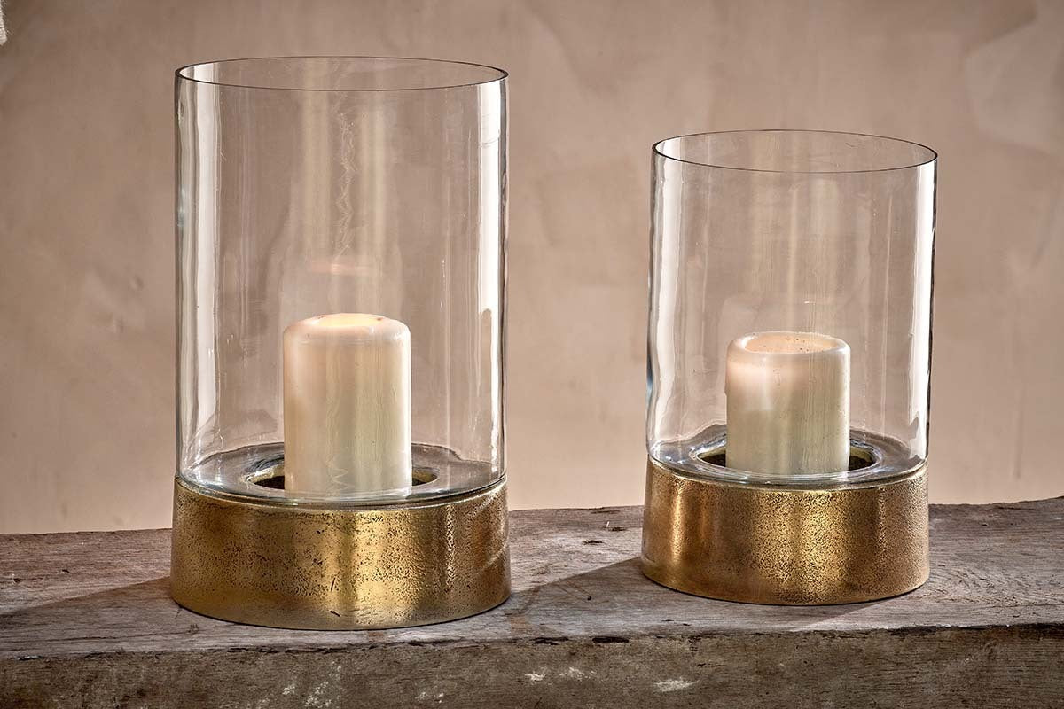 Two Rajura Hurricane Lanterns by nkuku, featuring gold metal bases, rest on a rustic wooden surface. Each glass holder has a white pillar candle, casting soft light against a neutral background that accentuates their elegant simplicity.