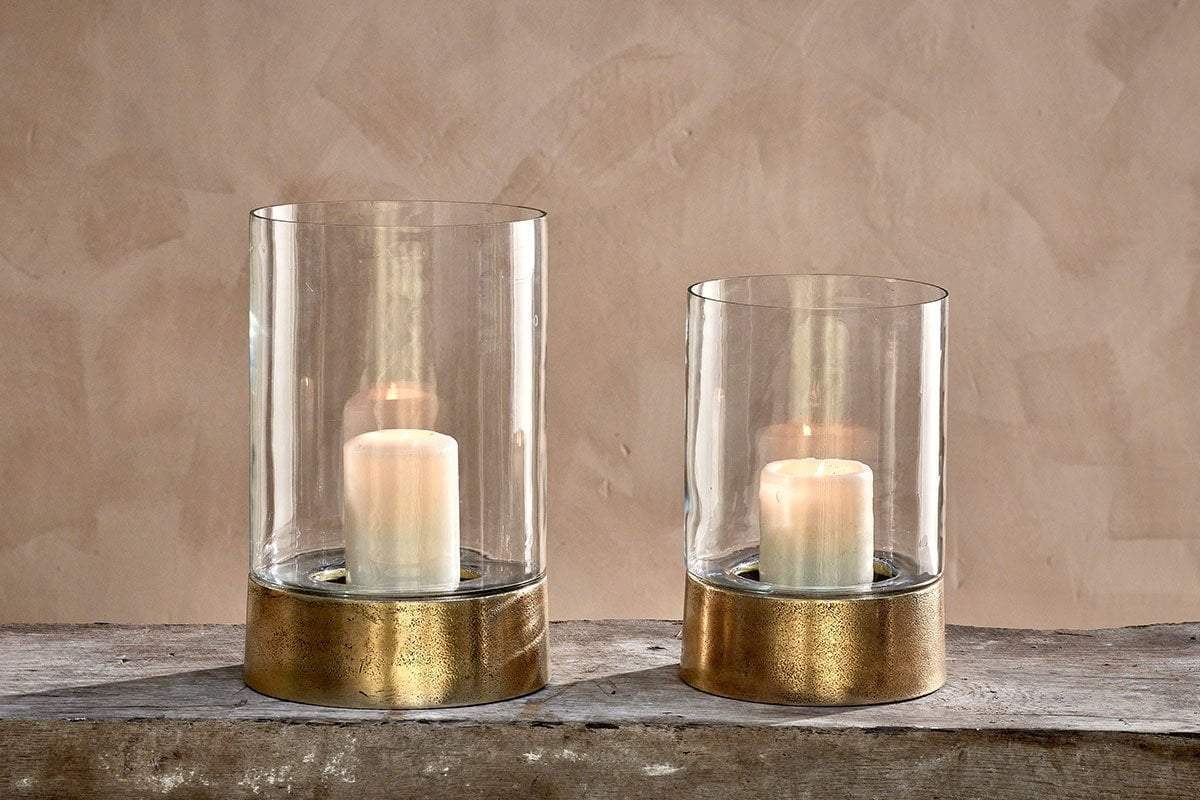 Two Rajura Hurricane Lanterns by nkuku, featuring white pillar candles and metallic gold bases, sit on a rustic wooden surface against a beige wall. The lanterns are different heights, ideal for adding ambient light to any room.