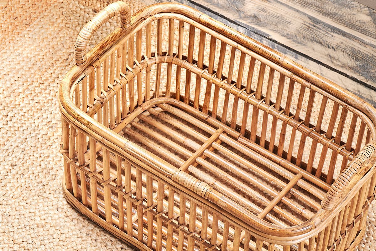 The nkuku Rammi Rattan Laundry Basket features an open design, a single handle, and natural rattan slats. Stylishly displayed on a woven mat by a rustic wooden wall, it offers attractive and practical storage.