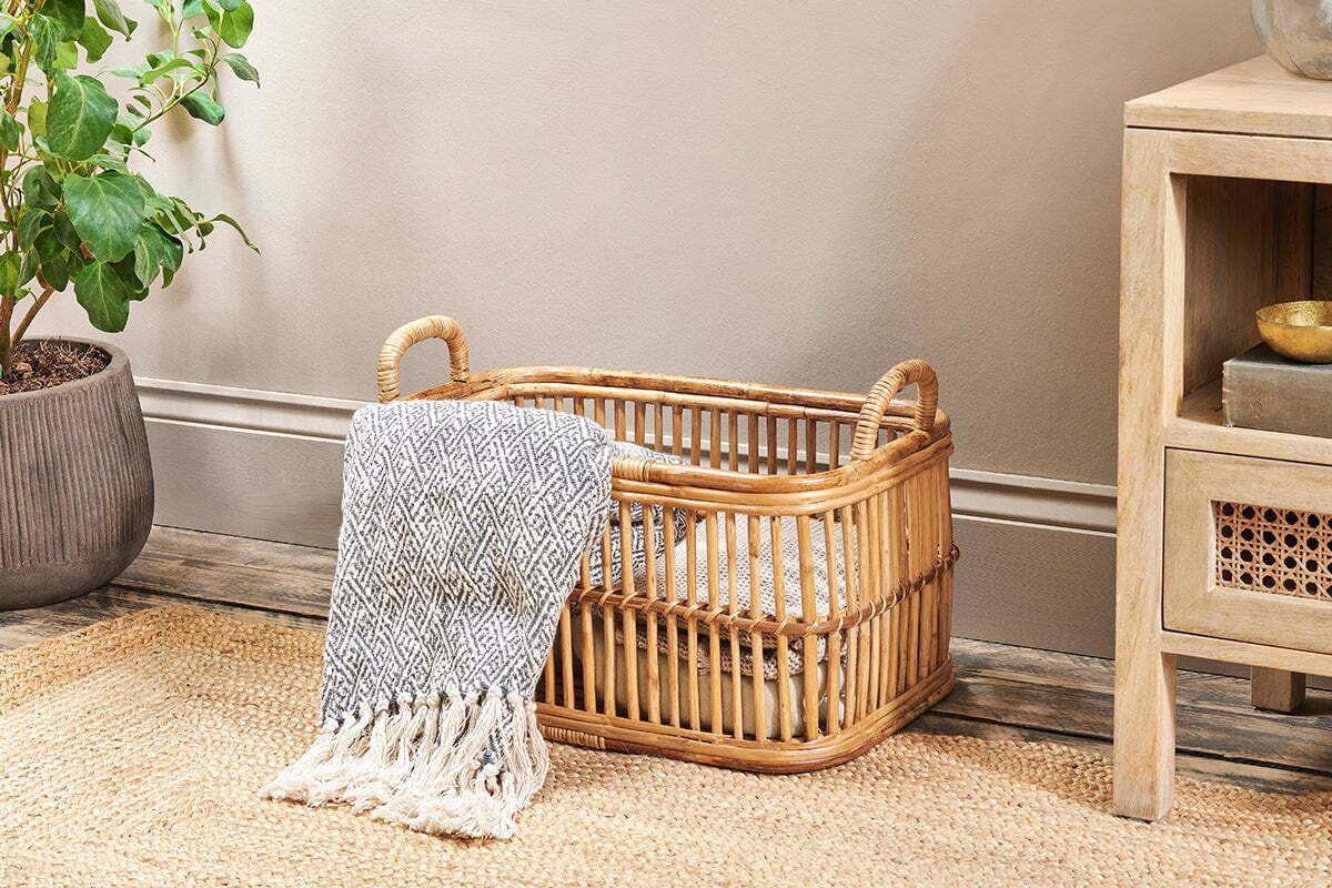 The nkuku Rammi Rattan Laundry Basket with two handles sits on a woven rug, draped with a gray and white fringed blanket. Nearby are a potted plant and a light wood cabinet with a gold bowl on its shelf.