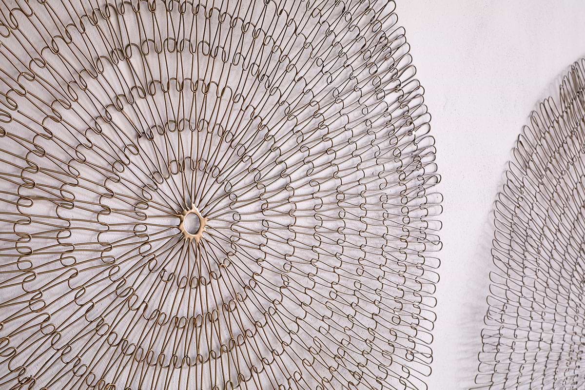 Close-up of the nkuku Rayan Wire Wall Art in Antique Brass, showcasing two large, circular sculptures with concentric rings and lacy geometric patterns. Only part of the second piece is visible on a light-colored wall.