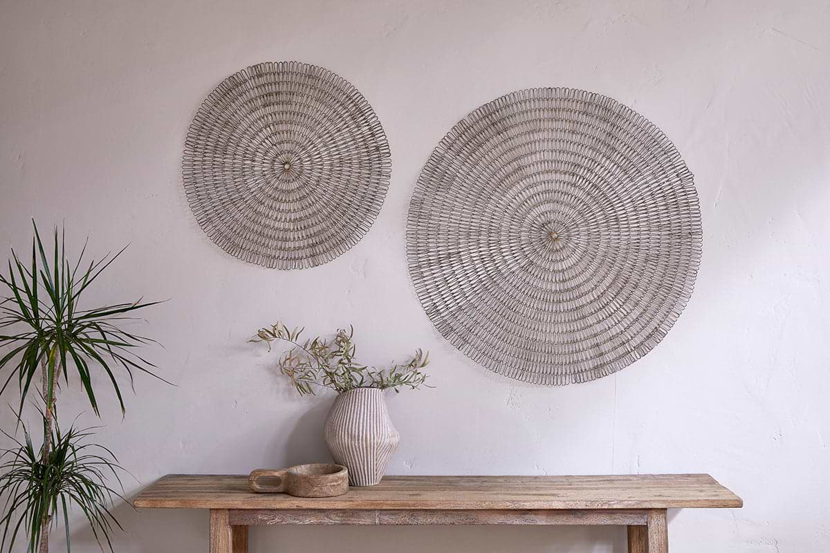 A minimalist interior showcases two large nkuku Rayan Wire Wall Art pieces in Antique Brass on a textured white wall, with a rustic wooden table below, accented by a striped ceramic vase, green branches, wooden bowl, and a potted spiky plant.