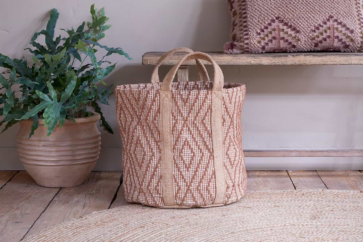 The Renu Jute Basket – Rust & Natural by nkuku sits on a wooden floor beside a leafy potted plant. Behind it is a wooden bench with a kilim cushion, and a round woven jute rug is partially visible in the foreground.