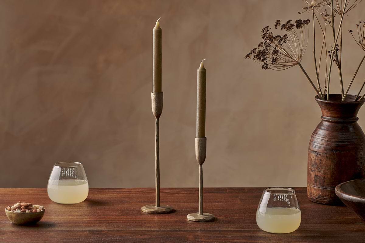 A wooden table with nkuku Rustic Soy Blend Dinner Candles in Olive Green (Set of 2), brass holders, two short glasses with pale yellow drinks, a bowl of nuts, and a rustic brown vase with dried plants against a warm brown backdrop.
