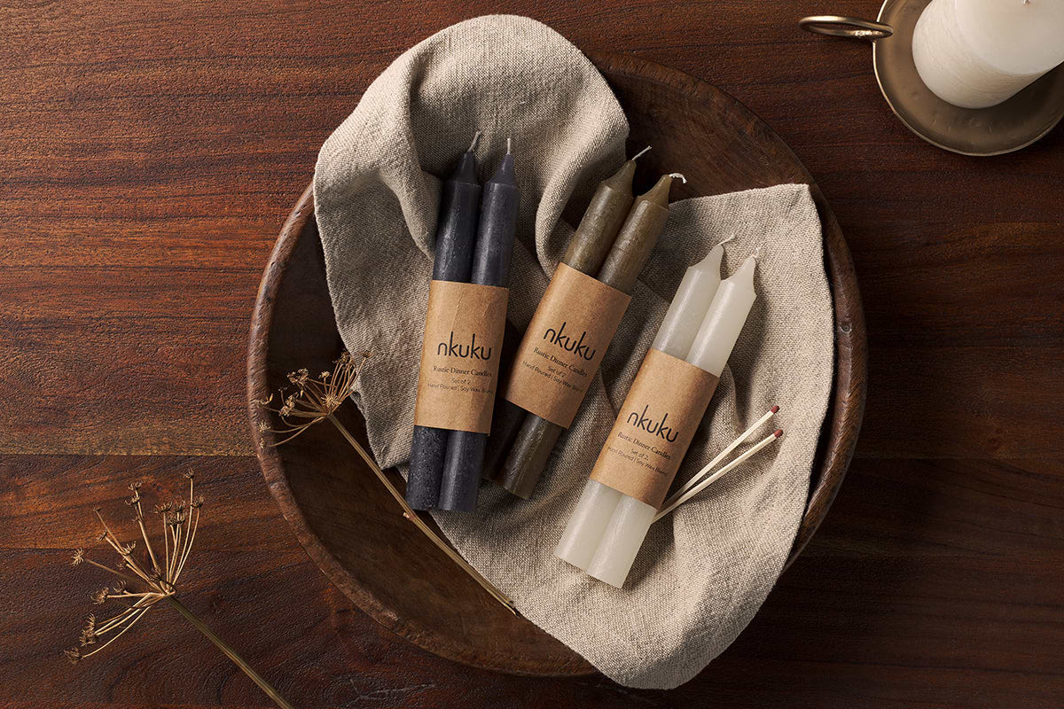 A round wooden bowl lined with beige cloth holds three bundles of nkuku Rustic Soy Blend Dinner Candles, including olive green (set of 2), each wrapped with brown nkuku labels. The bowl sits on dark wood next to dried plant stems.