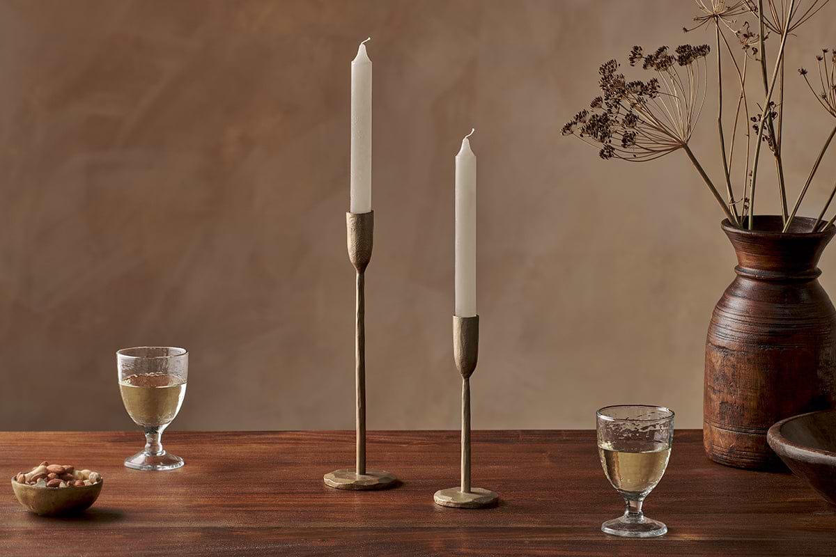 A wooden table displays two nkuku Rustic Soy Blend Dinner Candles in white, brass holders, wine glasses, a bowl of nuts, and a brown ceramic vase with dried flowers. A warm beige background adds to the cozy, elegant atmosphere.