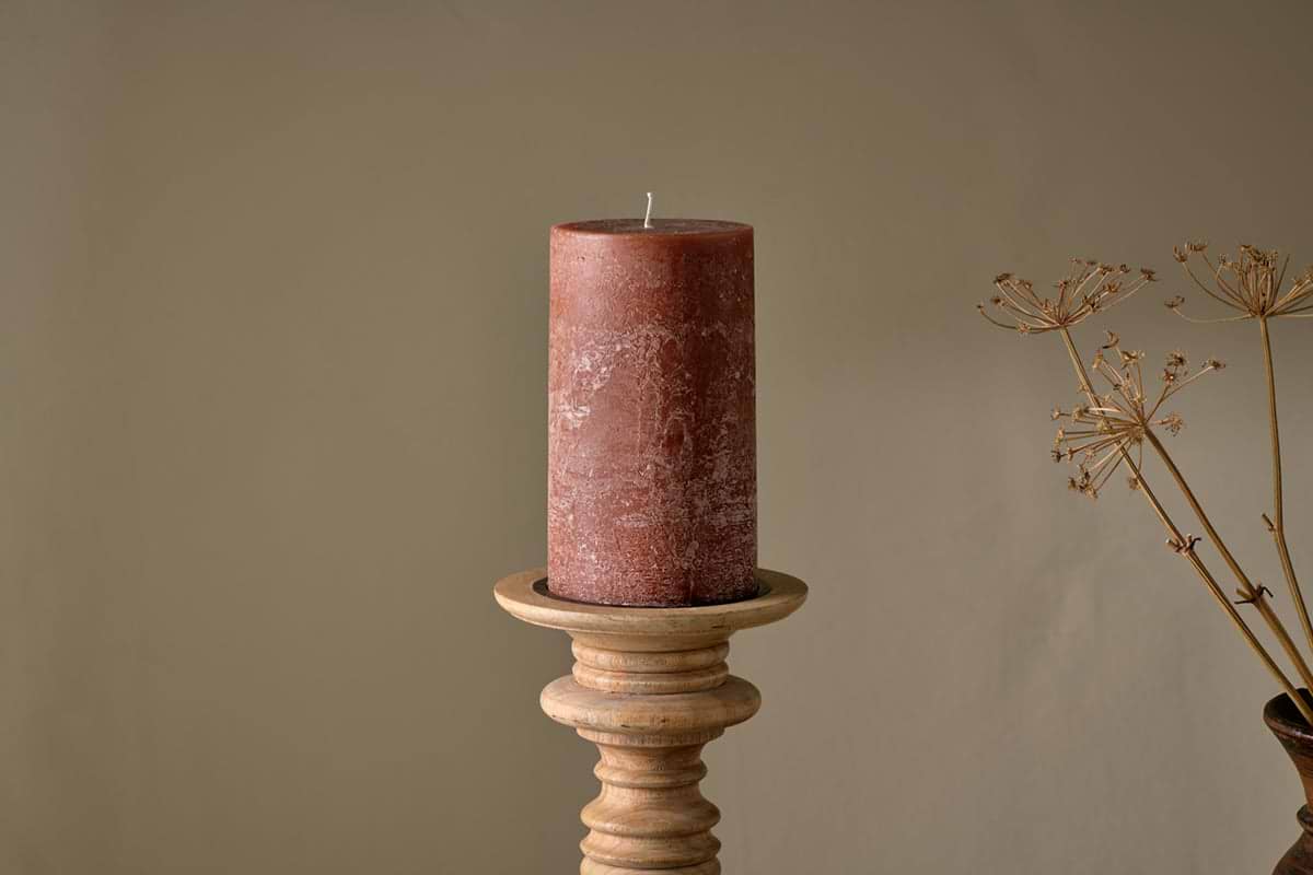 The nkuku Rustic Soy Blend Pillar Candle - Rust - Large, featuring a textured finish, is displayed on a wooden candlestick holder against a beige backdrop, with dried plant stems to the right for a natural accent.
