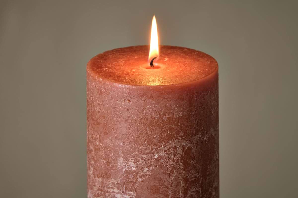 A close-up of the nkuku Rustic Soy Blend Pillar Candle in Rust (Large) showcases its textured reddish-brown wax, unique imperfections, and a warm flame glowing at the top against a softly blurred background.