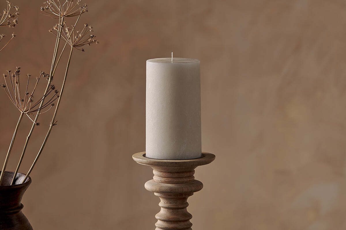 A large Rustic Soy Blend Pillar Candle in white by Nkuku sits on a wooden holder against a beige textured background, with delicate dried stems in a brown vase to the left for a natural, minimal aesthetic.