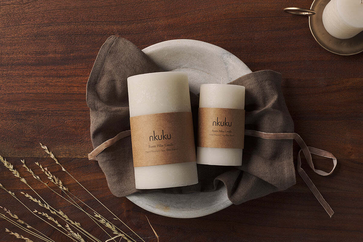 Two large white Rustic Soy Blend Pillar Candles by Nkuku with a textured finish rest on a gray stone plate atop a brown cloth on a wooden table. A third candle in a brass holder and dried grass complete the cozy, rustic scene.