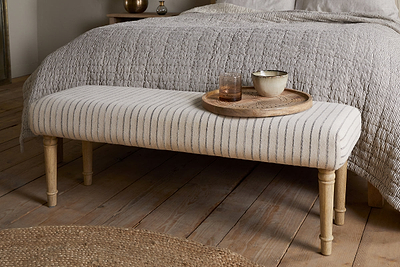 The nkuku Sanja Stripe Upholstered Bench - Grey with natural cotton fabric and wooden legs sits at the beds end. A round wooden tray holds a glass of water and bowl. The space features a textured quilt, woven rug, and wooden flooring.