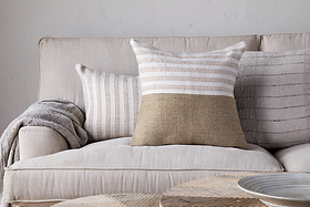 A beige sofa with a wool cushion cover and a folded gray blanket sits against a light wall. Two textured throw pillows, including the nkuku Samata Recycled Cushion Cover in Moss, add depth. A round woven coffee table is in the foreground.