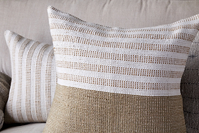 Two decorative throw pillows with woven textures, one in beige and white stripes and one with finer stripes, rest on a light sofa. Pair them with nkuku’s Samata Recycled Cushion Cover in Moss to enhance depth and style.