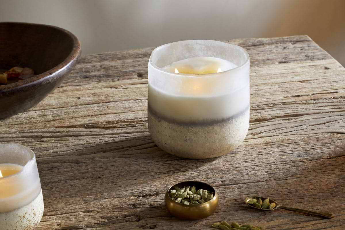 The nkuku Cardamom & Amber 3 Wick Scented Candle in frosted glass sits on a rustic wooden table next to a gold bowl of cardamom pods, with another nkuku candle and a wooden bowl adding sandalwood accents.