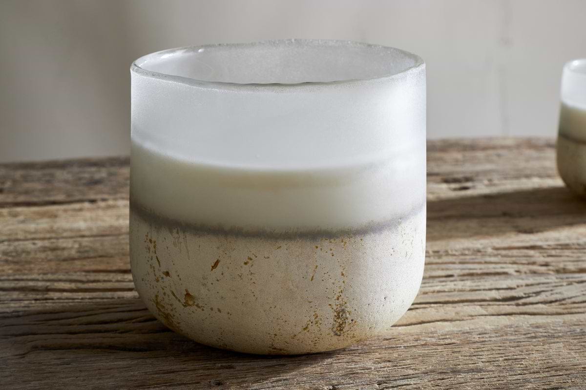 A frosted glass nkuku Cardamom & Amber 3 Wick Scented Candle with a white and beige gradient and rustic texture sits on a weathered wooden table.