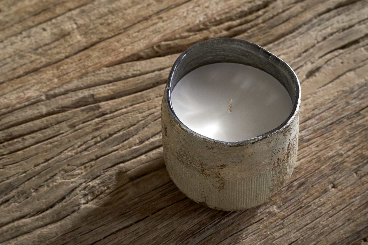 The Juniper & Fir 1 Wick Scented Candle by nkuku, housed in a rustic textured metal container, rests on a wooden surface and fills the room with its bespoke fragrance.