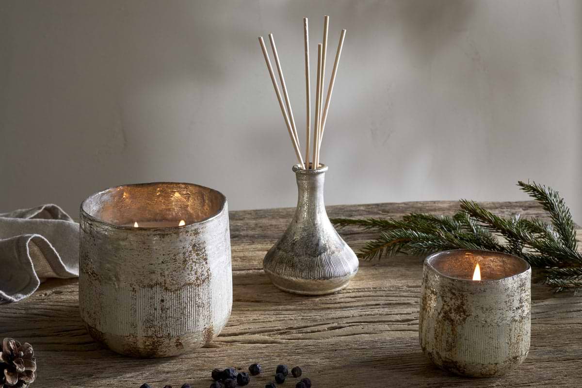 A rustic wooden table features the nkuku Juniper & Fir 1 Wick Scented Candle in a textured, reusable vessel, paired with two lit candles, a glass vase of reed diffuser sticks, pine branches, pinecones, and berries.