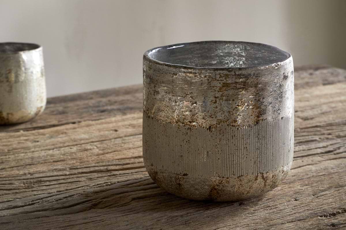 The nkuku Juniper & Fir 3 Wick Scented Candle rests on a rustic wooden table, its earthy ceramic vessel evoking the calm of winter; another similar candle is blurred behind, suggesting a serene, minimalist space.