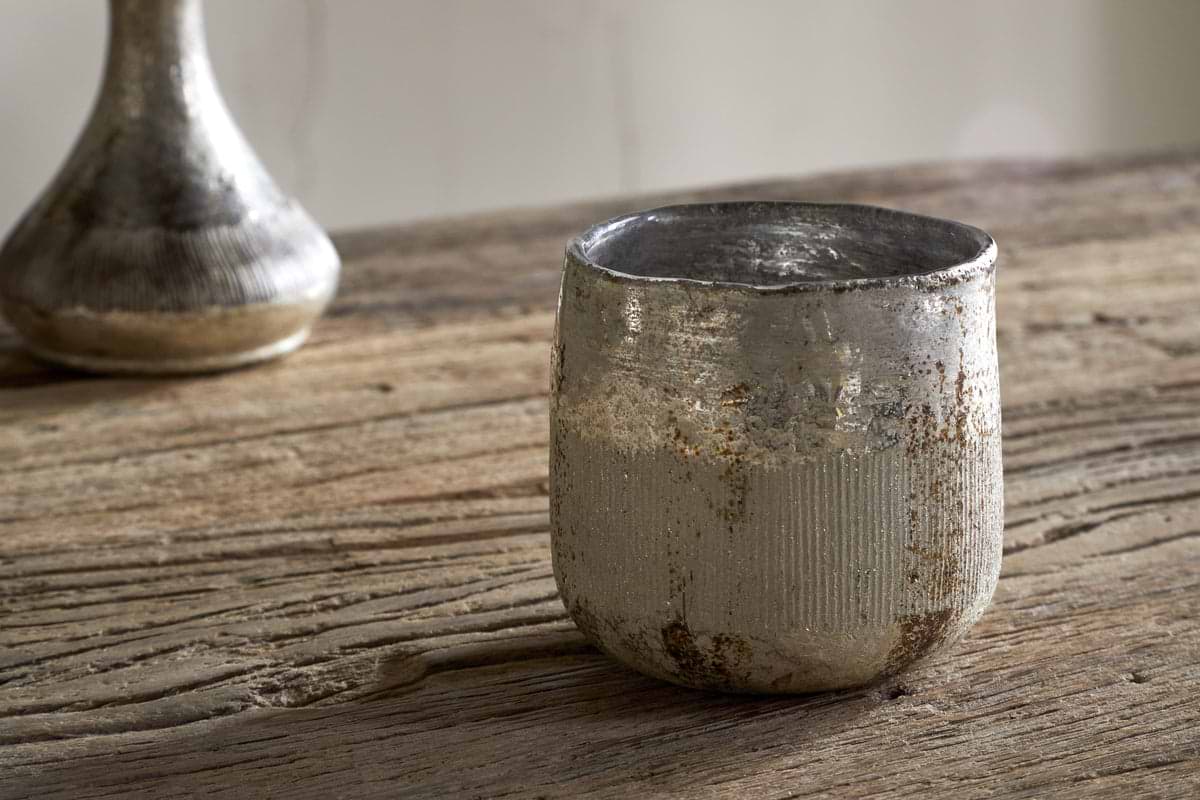 A nkuku Mandarin & Clove 1 Wick Scented Candle sits beside a weathered metallic cup on a rustic wooden table, with another blurred metallic object in the background.