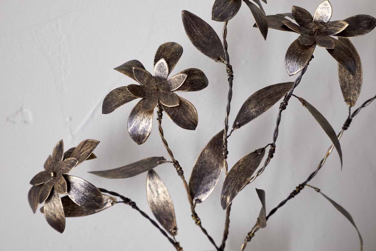 Close-up of the nkuku Svara Stem Flower, a metallic sculpture with stylized floral and leaf motifs, featuring an antique brass stem, set against a plain white background.