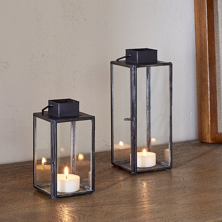 Two nkuku Sia Mini Table Lanterns in black, each holding a lit tealight, are arranged on a wooden surface beside a framed artwork and a glass jar of dried flowers.