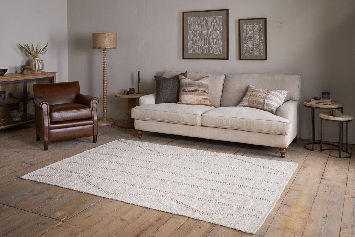 A cozy living room with a cream sofa, neutral pillows, a brown leather armchair, wooden side tables, and framed art on beige walls features the nkuku Shanti Wool & Cotton Rug - Natural on wood floors with warm, minimal decor.