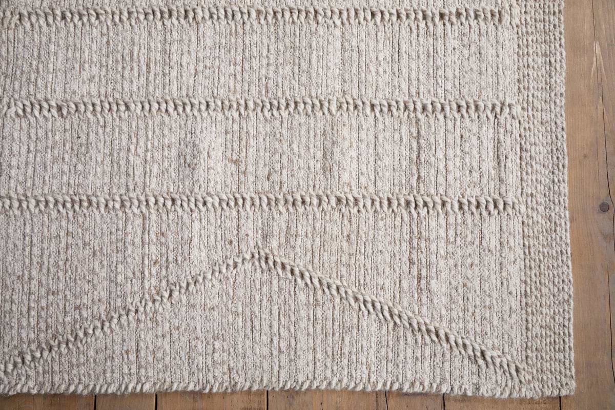 The Shanti Wool & Cotton Rug - Natural by nkuku showcases a textured beige weave with horizontal ridges and a triangle formed by diagonal lines in the corner, adding an elegant geometric touch to wooden floors.