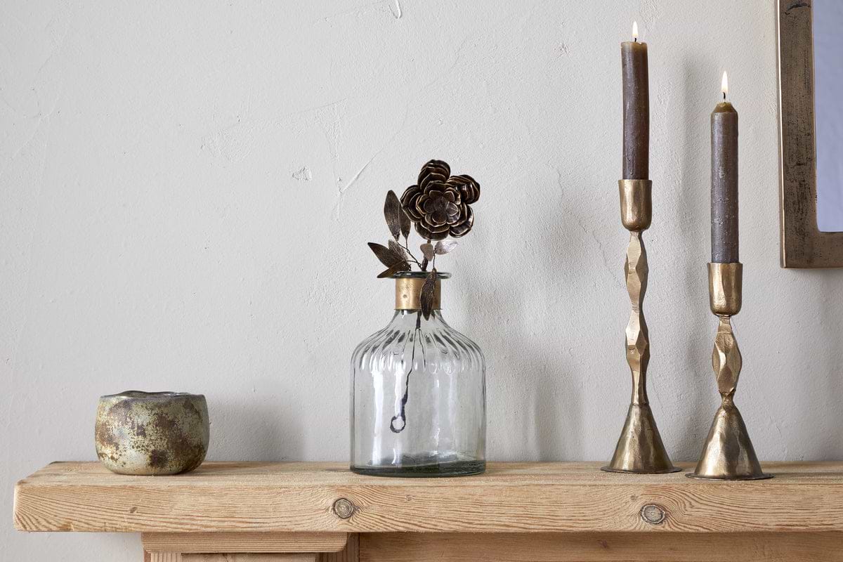 The nkuku Svara Stem Rose in antique brass, two lit brown candles in gold holders, and a small rustic cup rest on a wooden mantel against a light-colored wall.
