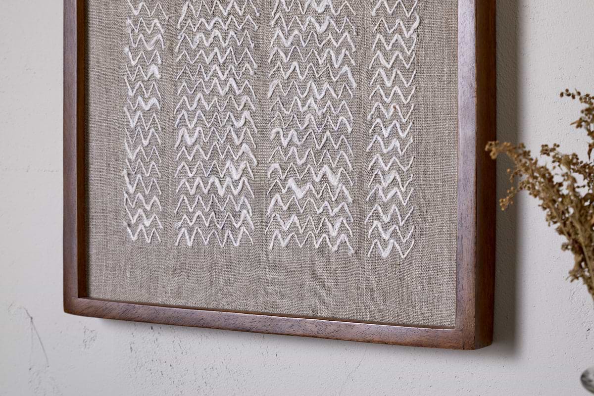 The Samaya Hand Stitched Wall Art - Natural by nkuku features white zigzag embroidery on beige linen, framed in warm brown sustainable mango wood—an elegant accent for your wall décor.