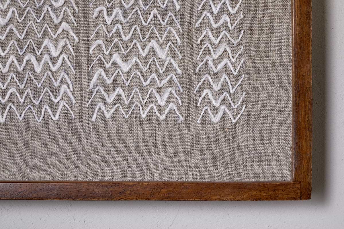 A close-up of the Samaya Hand Stitched Wall Art - Natural by nkuku, showing hand-stitched white zigzags on beige fabric in a sustainable mango wood frame, with the frame’s lower right corner and wall edge visible.