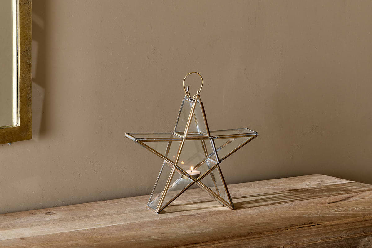 The Sanwi Standing Star Brass by nkuku, crafted from glass and metal with an antique brass finish, holds a small candle on a rustic wooden surface as sunlight from a nearby window casts a soft, warm glow over the minimalist scene.