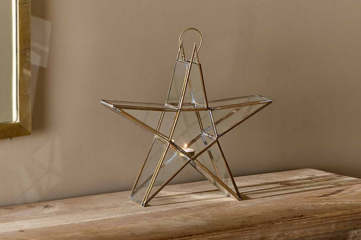 The nkuku Sanwi Standing Star Brass lantern, made of gold metal and glass, sits on a light wooden surface against a beige wall. Sunlight from the left highlights this festive piece and casts gentle shadows.
