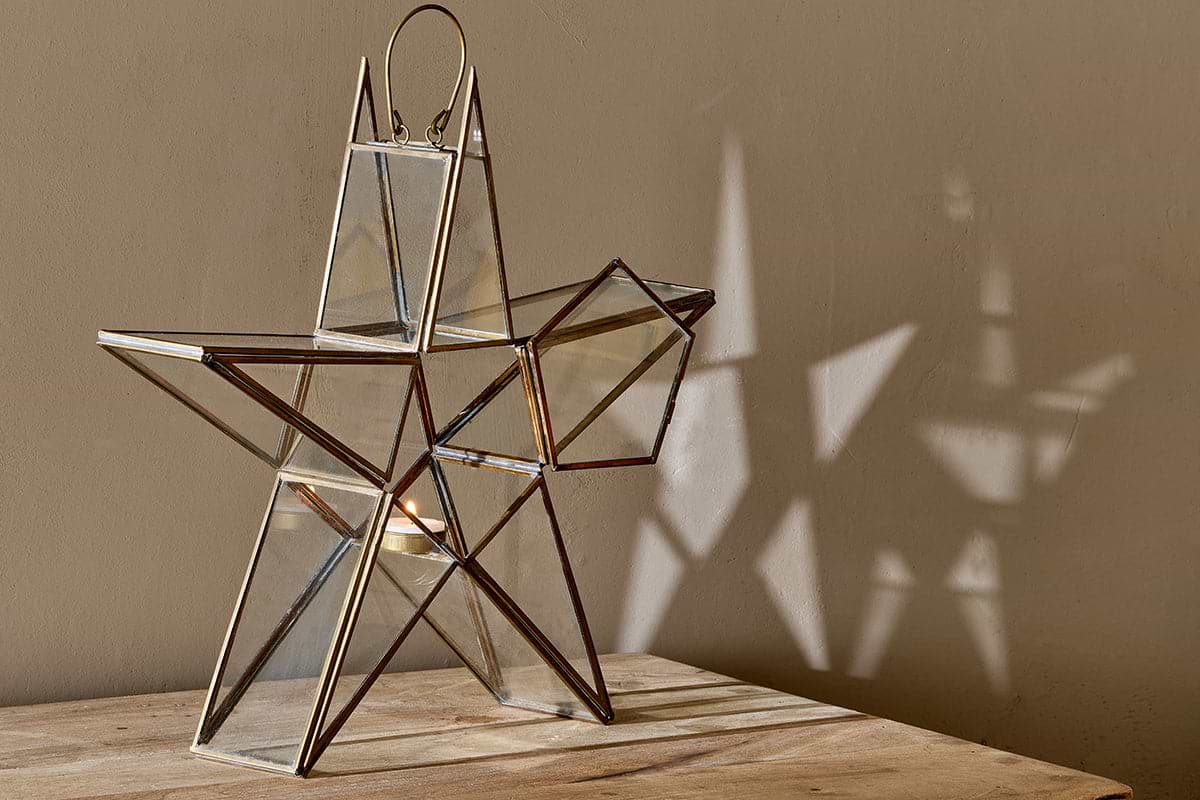 The Sanwi Standing Star Brass lantern by nkuku features glass and metal with brass-colored edges. Perfect for a small candle, it casts geometric shadows on a beige wall, bringing festive charm and elegance to any wooden surface.