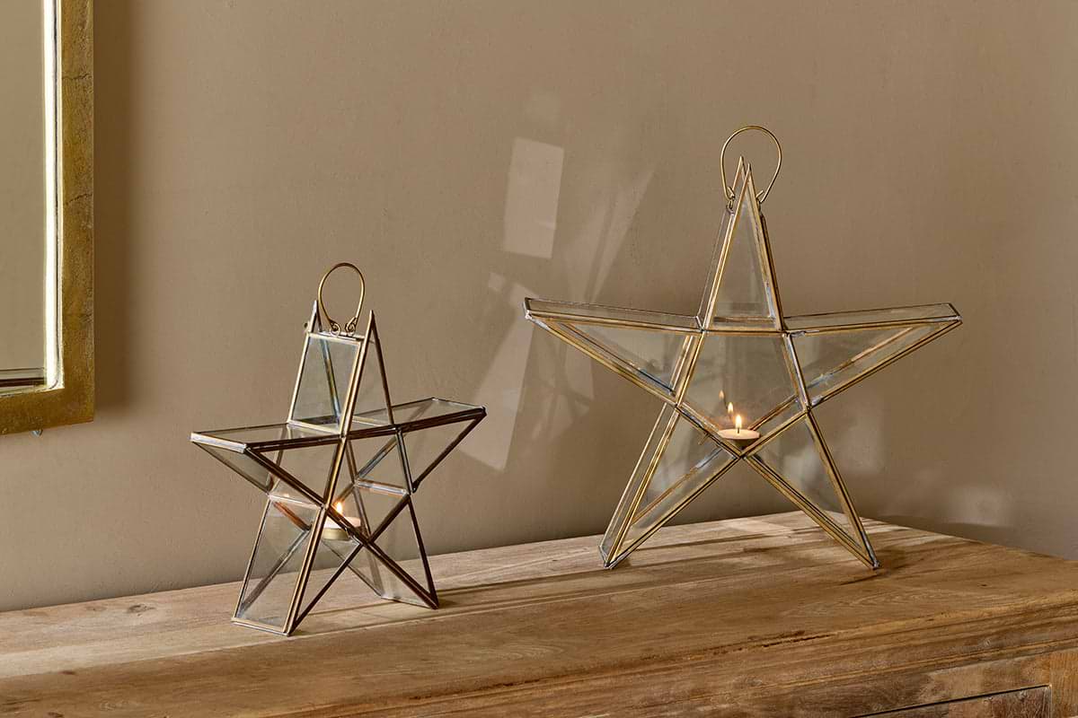 Two Sanwi Standing Star Brass lanterns by nkuku, each holding a lit tealight, rest on a wooden surface against a beige wall. Their glass and brass frames cast a soft glow. A partial framed mirror is visible on the left side.