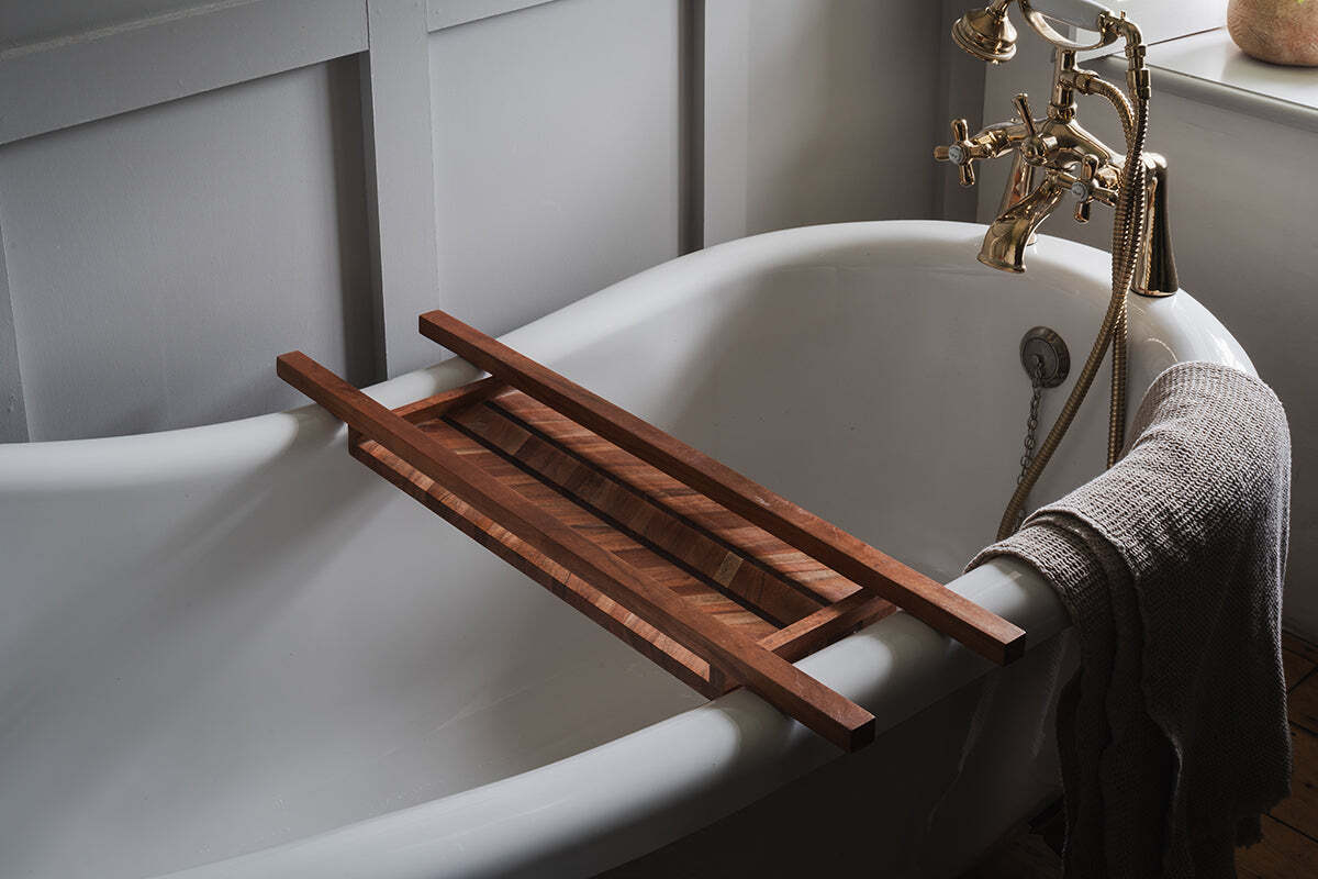 A white clawfoot bathtub features the nkuku Sukha Mango Bath Bridge - Natural, with brass fixtures and a textured gray towel draped over the edge, creating a serene, spa-like bathroom ambiance in gentle lighting.