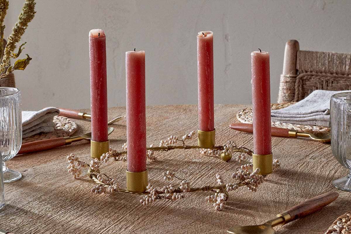 A mango wood table is set with glassware, napkins, and cutlery. At the center, nkuku’s Tanu Mango Wood Beaded Candle Holder in brass & natural displays four pink candles and white berries for an elegant festive touch.