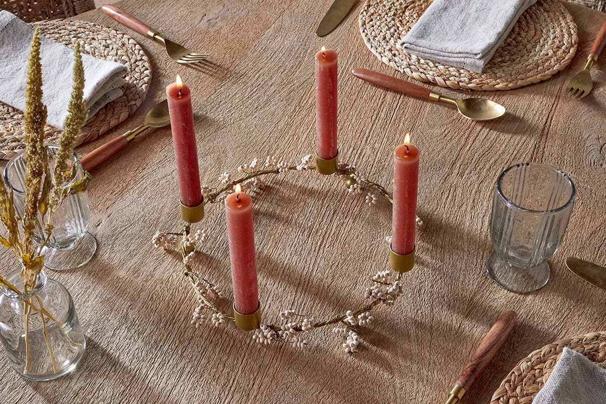 A mango wood table set for a meal features four nkuku Tanu Mango Wood Beaded Candle Holders in brass and natural, each topped with a red candle and surrounded by small white flowers. Place settings include woven placemats, gray napkins, gold utensils, and clear glasses.