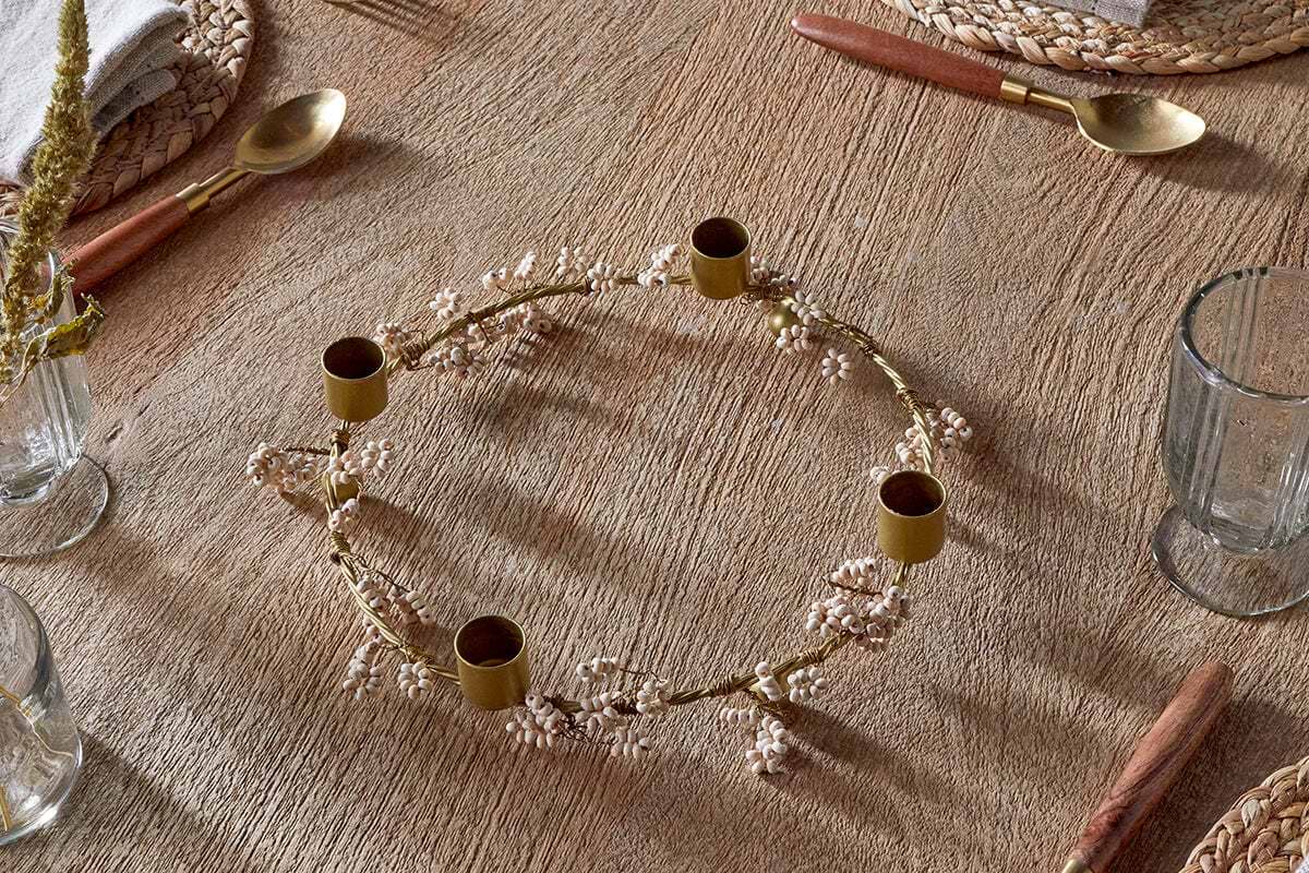 The nkuku Tanu Mango Wood Beaded Candle Holder in Brass & Natural sits on a rustic wooden table with woven placemats, mango wood and gold cutlery, clear glasses, and a beige napkin—ideal for softly lit festive tablescapes.