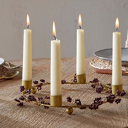 The Tanu Mango Wood Beaded Candle Holder in Brass & Ruby by nkuku, adorned with small purple berries and four tall cream candles, serves as an elegant centerpiece on a textured table set with plates, woven placemats, clear glasses, and gold utensils.